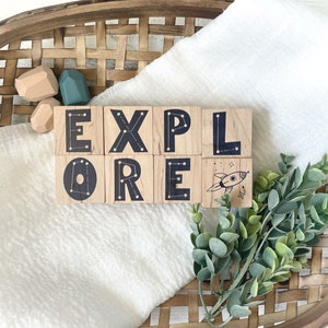 May include: Wooden blocks spelling out the word "EXPLORE" with a rocket ship design on the last block. The blocks have a constellation design on each side. The blocks are sitting on a white blanket with a woven basket in the background.