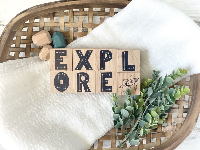 May include: Wooden blocks spelling out the word "EXPLORE" with a rocket ship design on the last block. The blocks have a constellation design on each side. The blocks are sitting on a white blanket with a woven basket in the background.