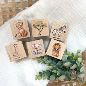 May include: Six wooden blocks with painted images of animals and a tree. The animals include a tiger, zebra, giraffe, elephant, and lion. The blocks are a light brown color and have a smooth, polished finish.
