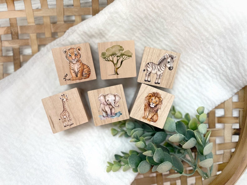 May include: Six wooden blocks with painted images of animals and a tree. The animals include a tiger, zebra, giraffe, elephant, and lion. The blocks are a light brown color and have a smooth, polished finish.