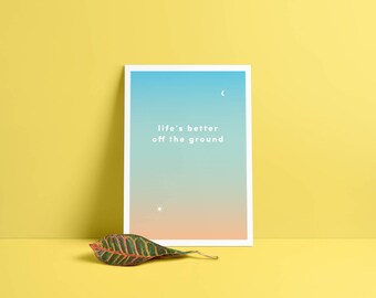 Puede incluir: Una impresión enmarcada en blanco con un fondo degradado azul y naranja. El texto "life's better off the ground" está impreso en blanco en la impresión.