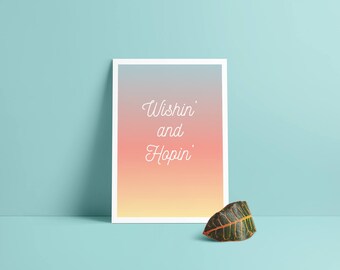 Puede incluir: Una impresión enmarcada en blanco con un fondo degradado de rosa, naranja y amarillo. El texto "Wishin' and Hopin'" está escrito en letra cursiva blanca.