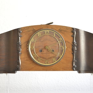 Könnte beinhalten: Eine antike Holz-Kaminuhr mit einem runden, goldenen Zifferblatt und schwarzen Zeigern. Die Uhr hat eine dunkelbraune Holzoptik und verzierte Metallapplikationen.