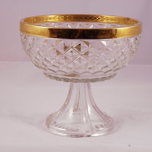 Puede incluir: Un cuenco de cristal transparente con un borde dorado. El cuenco tiene un patrón de diamante y se asienta sobre una base de pedestal.