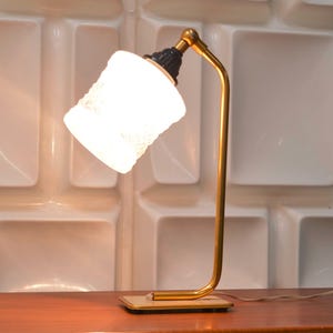Peut inclure: Une lampe de bureau vintage avec une structure en métal doré et un abat-jour en verre blanc texturé. L'abat-jour est incliné et émet une lumière douce. La lampe repose sur une base rectangulaire et a un accent noir en haut.