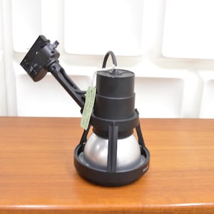 May include: A black spotlight with a silver bulb, mounted on a black arm. The spotlight sits on a wooden surface. A green tag hangs from the top of the spotlight.