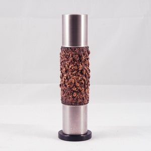Könnte beinhalten: Eine silberne Vase mit einem strukturierten Kupferband um die Mitte. Die Vase hat einen schwarzen Sockel.