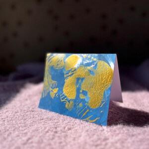May include: A folded greeting card with a blue and gold abstract design. The card is standing upright on a pink surface, with the interior of the card visible. The design features gold paint strokes on a blue background.