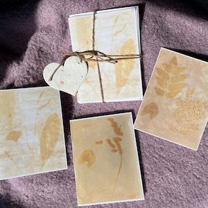 May include: A set of botanical art cards with pressed leaves and plant details in beige and brown. The cards are tied with twine and have a heart-shaped tag. The cards are displayed on a purple surface.