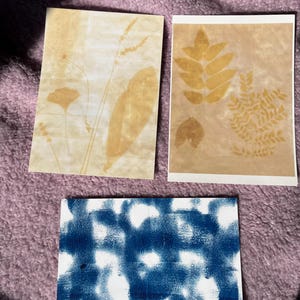 May include: Three handmade art prints. Two prints feature botanical impressions in shades of tan and brown on textured paper. The third print has a blue and white abstract design. The prints are displayed on a purple surface.