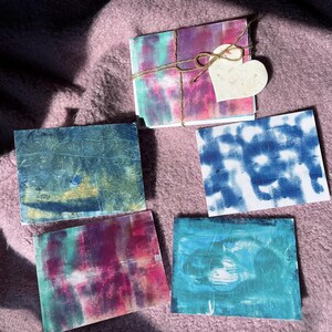 May include: A set of handmade greeting cards with abstract watercolour designs in blue, pink, and green hues. The cards are tied with twine and a heart-shaped tag. The cards are displayed on a pink, textured surface.