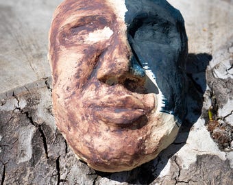 May include: A ceramic face mask with a split design. One half is brown, the other is black and white. The mask is resting on a weathered, cracked wooden surface, with a blurred green background.