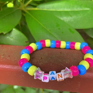 May include: A colorful beaded bracelet with the letters "PAN" spelled out in white letter beads. The bracelet features pink, yellow, and blue beads. The letters are on clear star-shaped beads. The bracelet is on a brown surface with green leaves in the background.