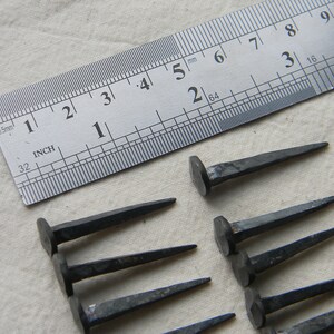 Puede incluir: Primer plano de diez clavos de hierro forjados a mano, con una regla que mide 1 pulgada (2,5 cm) de largo. Los clavos tienen una cabeza cuadrada y una punta puntiaguda.
