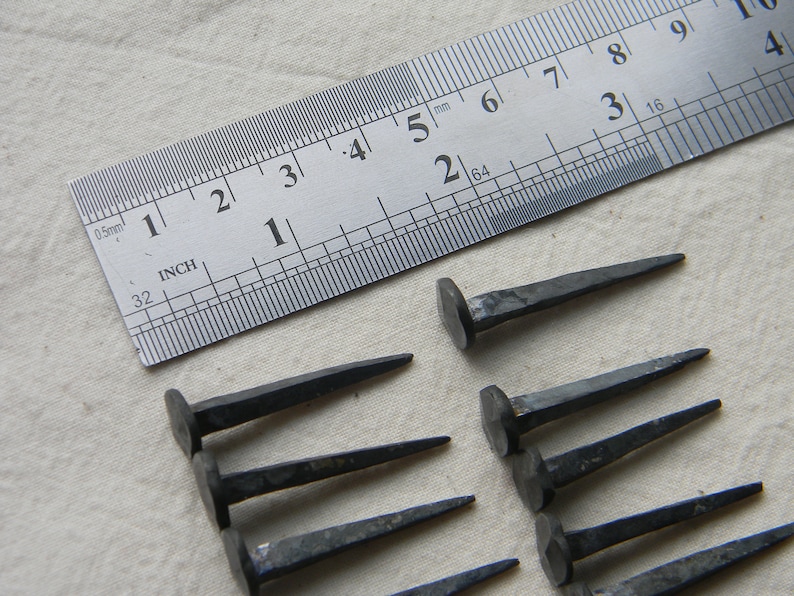 Puede incluir: Primer plano de diez clavos de hierro forjados a mano, con una regla que mide 1 pulgada (2,5 cm) de largo. Los clavos tienen una cabeza cuadrada y una punta puntiaguda.