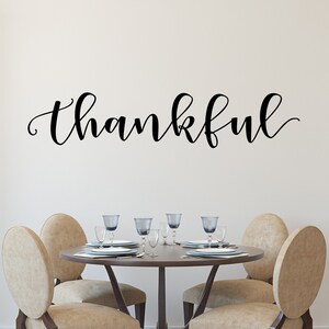May include: A dining room with a round wooden table and four beige chairs. The word "thankful" is written in black script on the wall. The table is set with plates, glasses, and cutlery. The room has a neutral colour scheme and a rug on the floor.