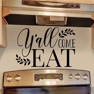 May include: Kitchen wall decor with the phrase "Y'all Come Eat" in black script and block letters, accented with leaf designs. The text is above a stainless steel stove and range hood, with light wood cabinets.