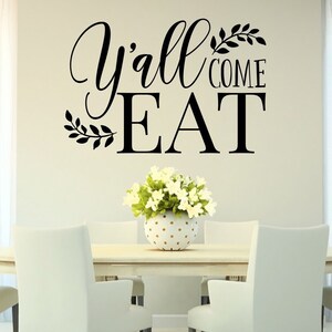 May include: A dining room scene featuring a light-colored oval table surrounded by six white chairs. A floral arrangement sits in the center. Above the table, the wall displays the text "Y'all COME EAT" in black script and block letters.