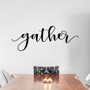 May include: A black cursive "gather" wall decal above a wooden dining table. The table is set with clear chairs and candles. The chair in the centre has a black and white patterned cushion.