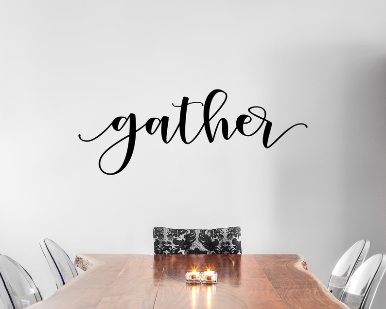May include: A black cursive "gather" wall decal above a wooden dining table. The table is set with clear chairs and candles. The chair in the centre has a black and white patterned cushion.