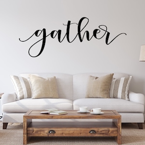 May include: A living room with a white sofa, beige and striped cushions, and a wooden coffee table. The word "gather" is written in black script on the wall. A floor lamp stands beside the sofa, creating a warm and inviting atmosphere.