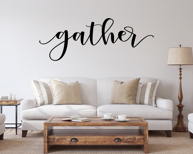 May include: A living room with a white sofa, beige and striped cushions, and a wooden coffee table. The word "gather" is written in black script on the wall. A floor lamp stands beside the sofa, creating a warm and inviting atmosphere.