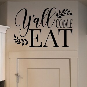 May include: Black wall decal with the words "Y'all come EAT" in a decorative script font. The text is accompanied by leaf accents. The decal is placed above a white cabinet.