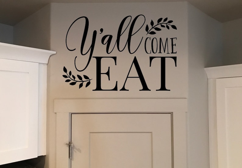 May include: Black wall decal with the words "Y'all come EAT" in a decorative script font. The text is accompanied by leaf accents. The decal is placed above a white cabinet.