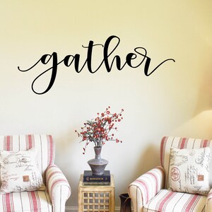 May include: A black script wall decal reads "gather". Two armchairs with red and white stripes and decorative cushions are positioned below. A small table with a vase of red berries and a floor lamp complete the inviting interior design.