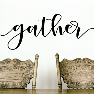 May include: A wall decoration featuring the word "gather" in black cursive script. Below the text are two intricately carved wooden chairs, likely for a dining room or kitchen, creating a welcoming atmosphere.