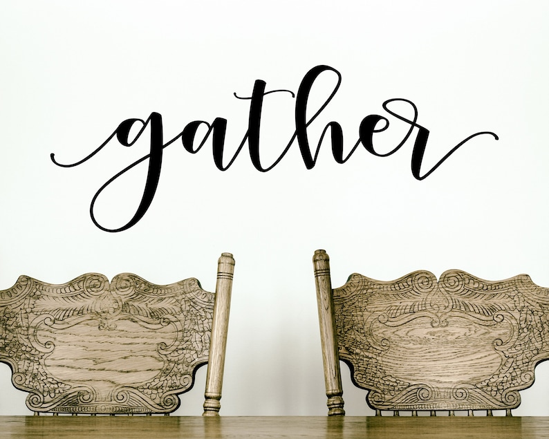 May include: A wall decoration featuring the word "gather" in black cursive script. Below the text are two intricately carved wooden chairs, likely for a dining room or kitchen, creating a welcoming atmosphere.