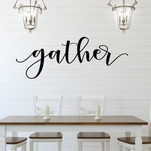 May include: A dining room setting with a wooden table and four white chairs. The word "gather" is written in black script on the white shiplap wall. Two candle holders are on the table, and two hanging light fixtures are above.