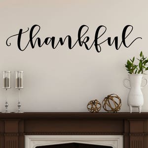 May include: A wall decoration with the word "thankful" in elegant black script. Below, a dark brown mantel displays decorative items: two candle holders, a white vase with greenery, and two decorative spheres. The wall is painted a light beige.