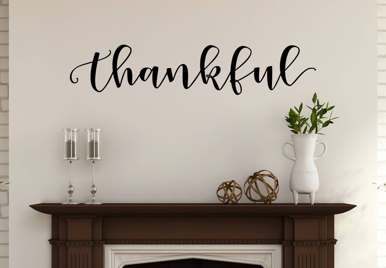May include: A wall decoration with the word "thankful" in elegant black script. Below, a dark brown mantel displays decorative items: two candle holders, a white vase with greenery, and two decorative spheres. The wall is painted a light beige.