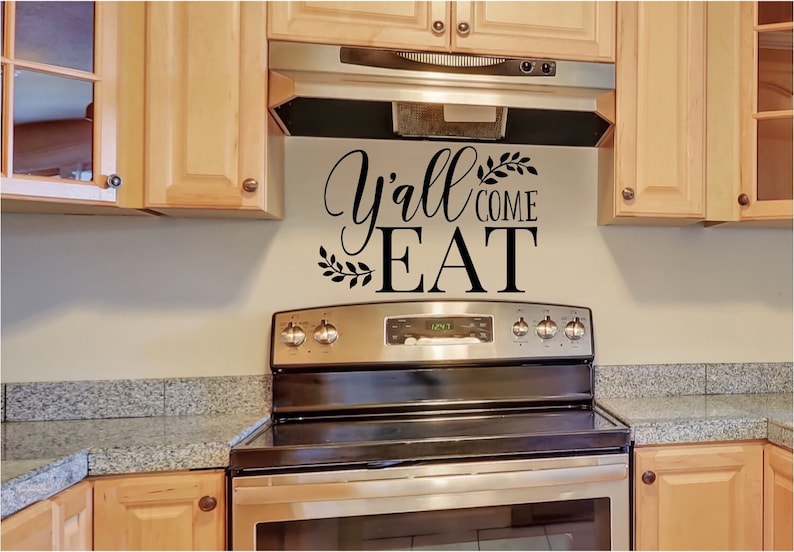 May include: Kitchen wall decor with the phrase "Y'all Come Eat" in black script and block letters, accented with leaf designs. The text is above a stainless steel stove and range hood, with light wood cabinets.