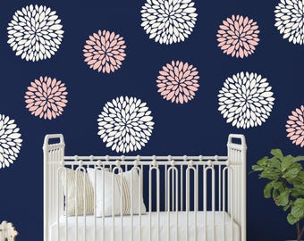 May include: A dark blue wall featuring a pattern of white and light pink dahlia flower wall decals. Below the wall, a white crib with vertical slats holds two pillows, one solid white and one with beige stripes, next to a green potted plant.