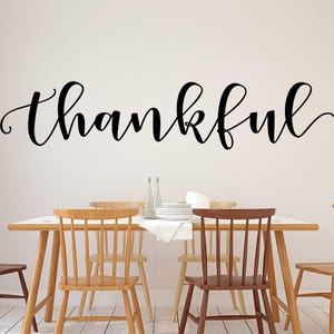 May include: A dining room scene with a wooden table and chairs. The word "thankful" is written in elegant black script on the wall above the table. The chairs are various shades of brown and tan.