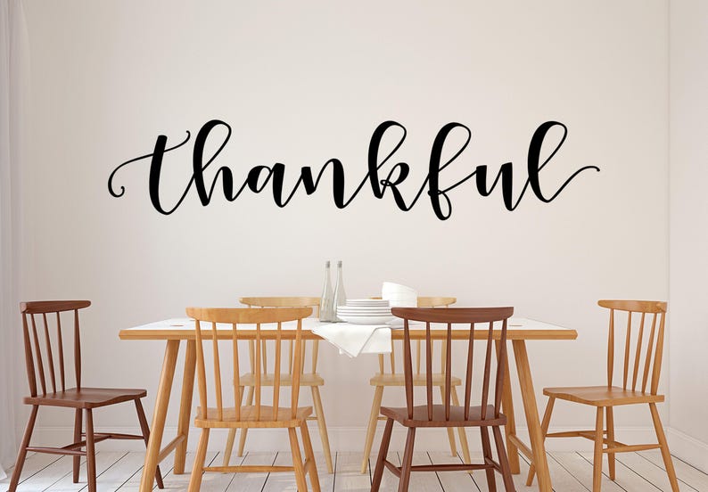 May include: A dining room scene with a wooden table and chairs. The word "thankful" is written in elegant black script on the wall above the table. The chairs are various shades of brown and tan.