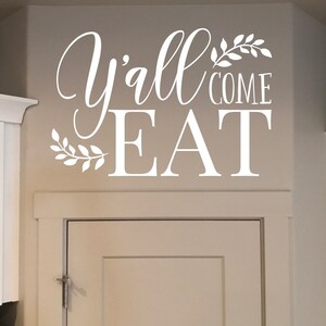 May include: White wall decal with the words "Y'all Come Eat" in a decorative script font. The text is accompanied by leaf accents. The decal is applied above a white cabinet door.