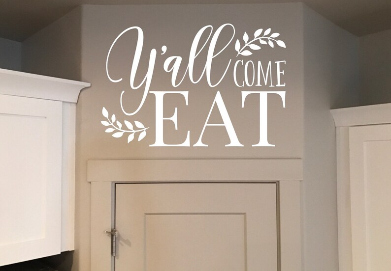 May include: White wall decal with the words "Y'all Come Eat" in a decorative script font. The text is accompanied by leaf accents. The decal is applied above a white cabinet door.