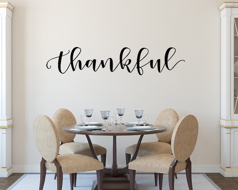 May include: A dining room with a round wooden table and four beige chairs. The word "thankful" is written in black script on the wall. The table is set with plates, glasses, and cutlery. The room has a neutral colour scheme and a rug on the floor.