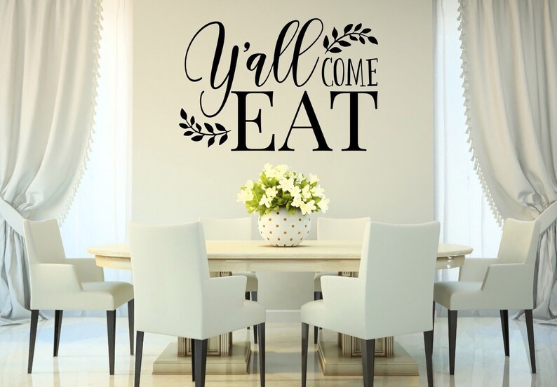 May include: A dining room scene featuring a light-colored oval table surrounded by six white chairs. A floral arrangement sits in the center. Above the table, the wall displays the text "Y'all COME EAT" in black script and block letters.