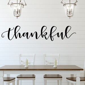 May include: A dining room setting with a white table and chairs, and a dark wood tabletop. The word "thankful" is written in elegant black script on the white wall above the table. Two candle holders are on the table, and two decorative hanging lights are above.