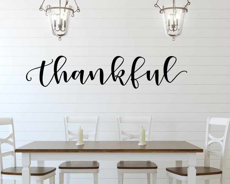 May include: A dining room setting with a white table and chairs, and a dark wood tabletop. The word "thankful" is written in elegant black script on the white wall above the table. Two candle holders are on the table, and two decorative hanging lights are above.