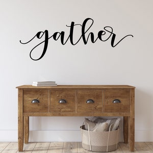 May include: A wooden console table with four drawers and black handles. The word "gather" is written in elegant black script above the table. A woven basket and books are also visible.