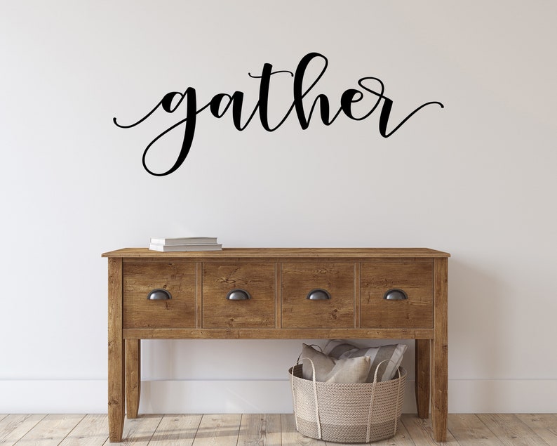 May include: A wooden console table with four drawers and black handles. The word "gather" is written in elegant black script above the table. A woven basket and books are also visible.