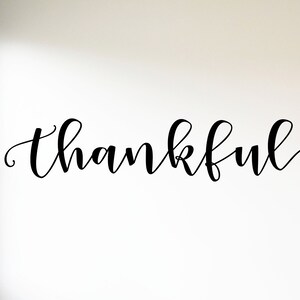 May include: A black cursive word "thankful" is displayed on a white wall. The script is elegant and flowing. The wall is plain white, and a window frame is visible on the left side of the image.