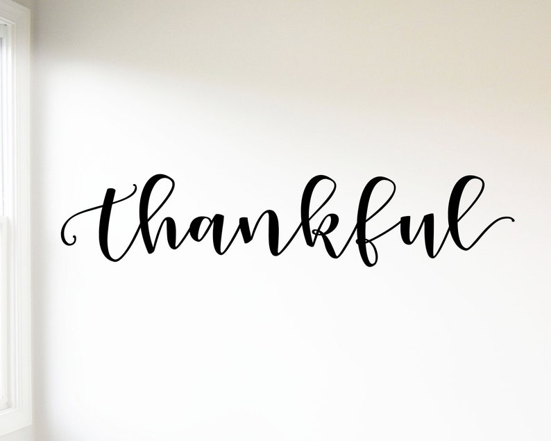 May include: A black cursive word "thankful" is displayed on a white wall. The script is elegant and flowing. The wall is plain white, and a window frame is visible on the left side of the image.