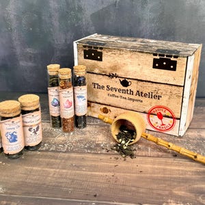 May include: A wooden box with the text "The Seventh Atelier" and several glass vials filled with tea. A wooden tea scoop with tea leaves is in front of the box. The vials have cork stoppers and labels with illustrations.