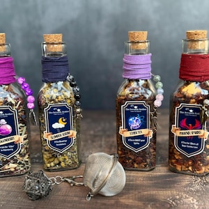 May include: Four clear glass bottles filled with loose leaf tea, each topped with a cork stopper and wrapped in coloured fabric. The labels read Dragonberry, Slumber Serum, Ether Tea, and Phoenix Sparks. A metal tea infuser sits in the foreground.